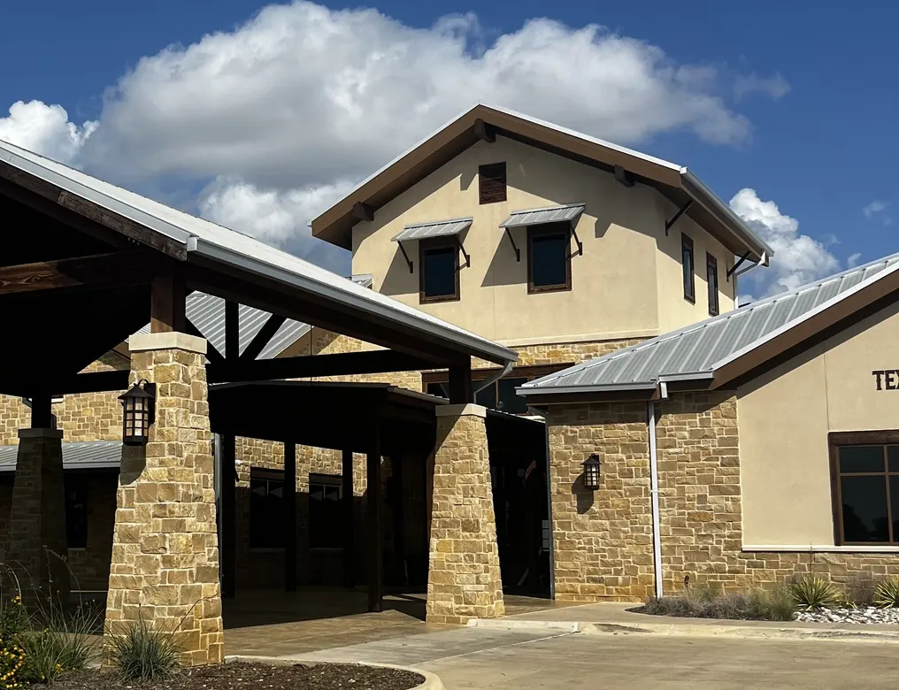 Stone and timber clubhouse entrance with a covered drive-up under blue sky at Texas Rangers Golf Club
