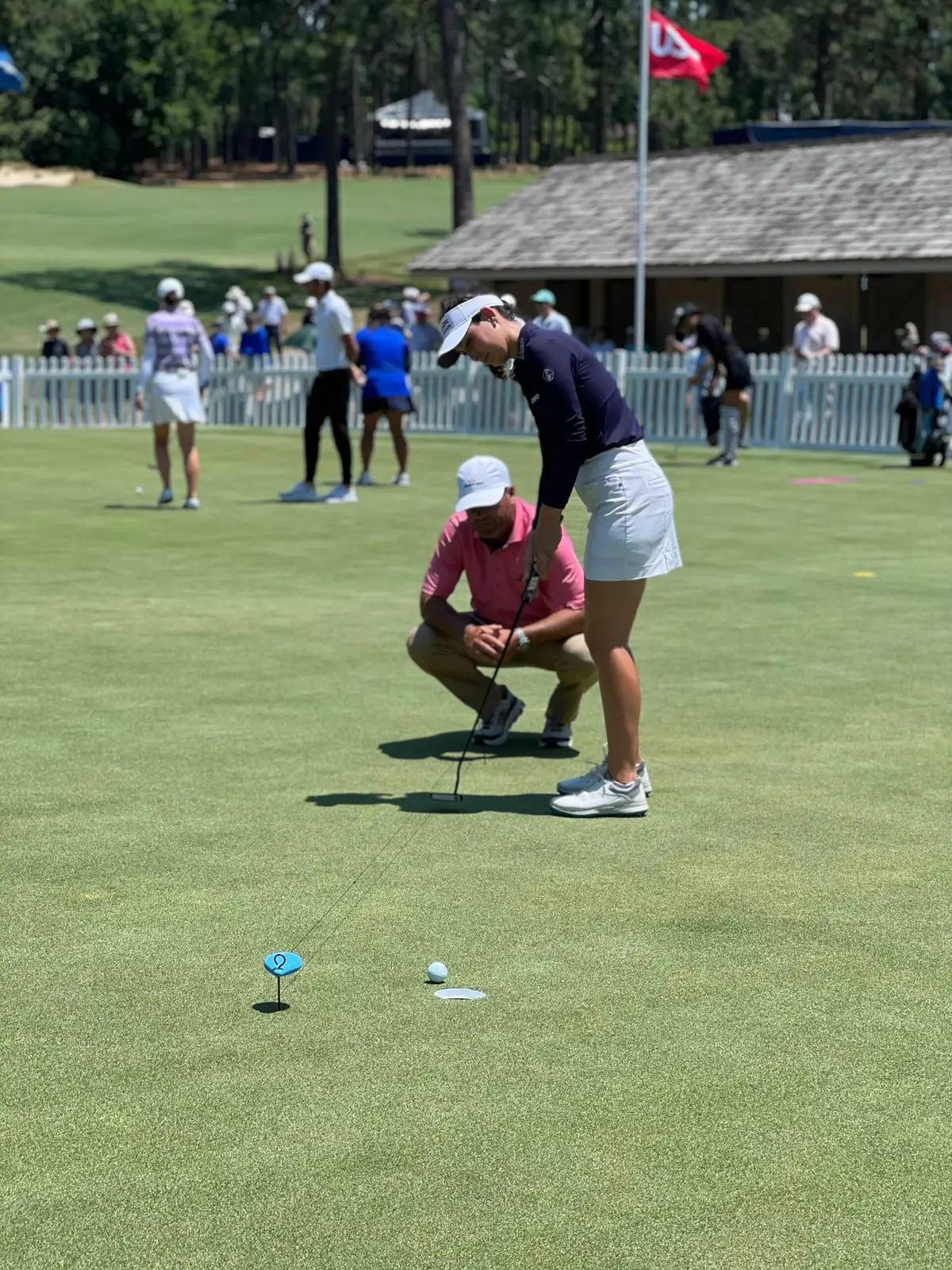 Joey Wuertemberger crouches behind a player putting on a practice green, with spectators along a white fence