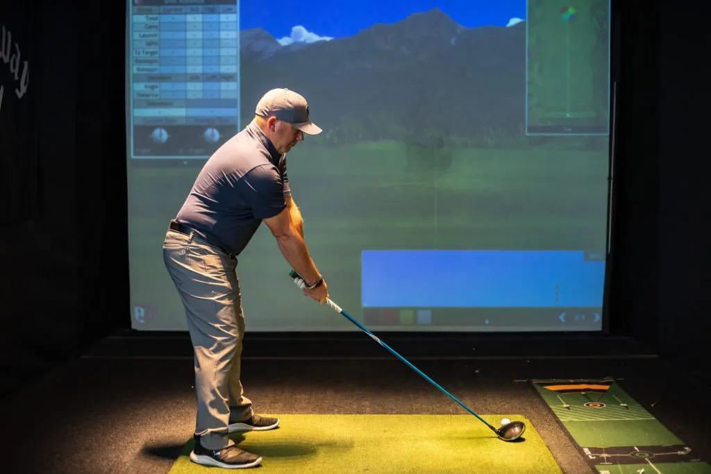 Denny Walters Jr. addressing a ball with a driver in an indoor simulator bay, a virtual fairway and shot data on the screen