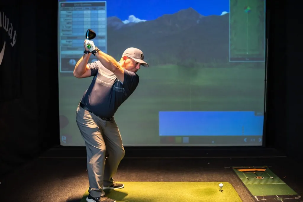 Denny Walters Jr. at the top of his backswing in an indoor simulator bay, a virtual fairway on the screen behind him
