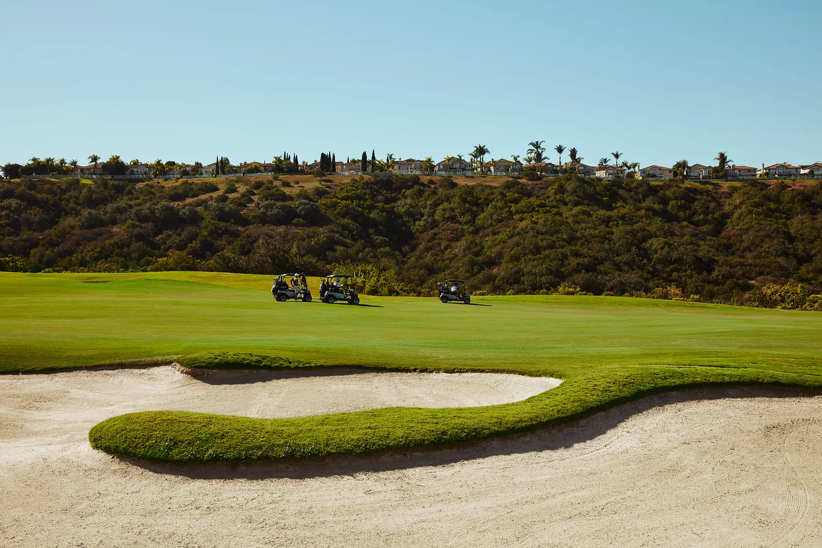 A curved sand bunker in front of a wide fairway with golf carts and hillside homes at The Grand
