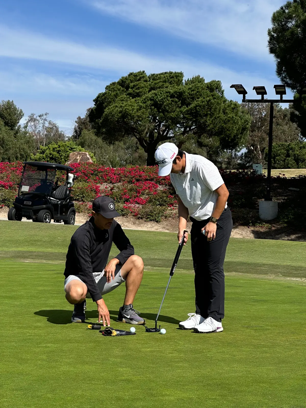 A coach crouches on a practice green beside a player lining up a putt, with flowering shrubs and a golf cart behind
