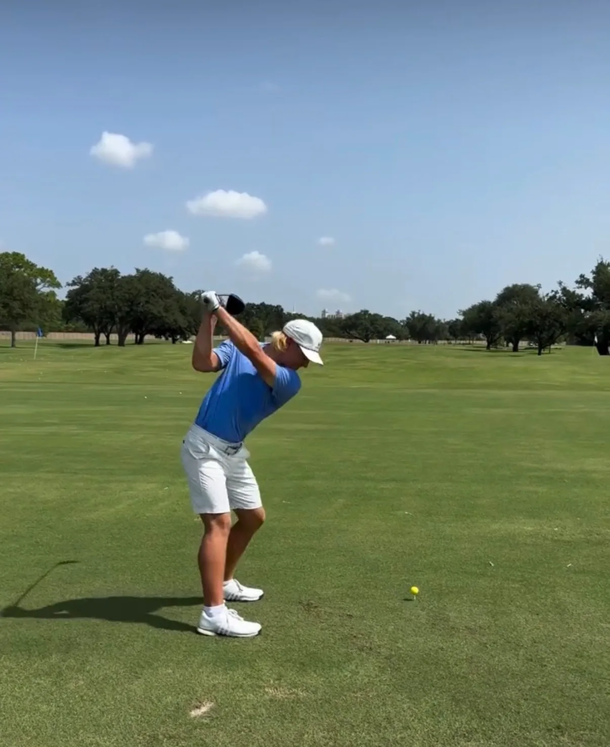 A golfer in a blue shirt at the top of the backswing on a grass practice tee, with oak trees behind