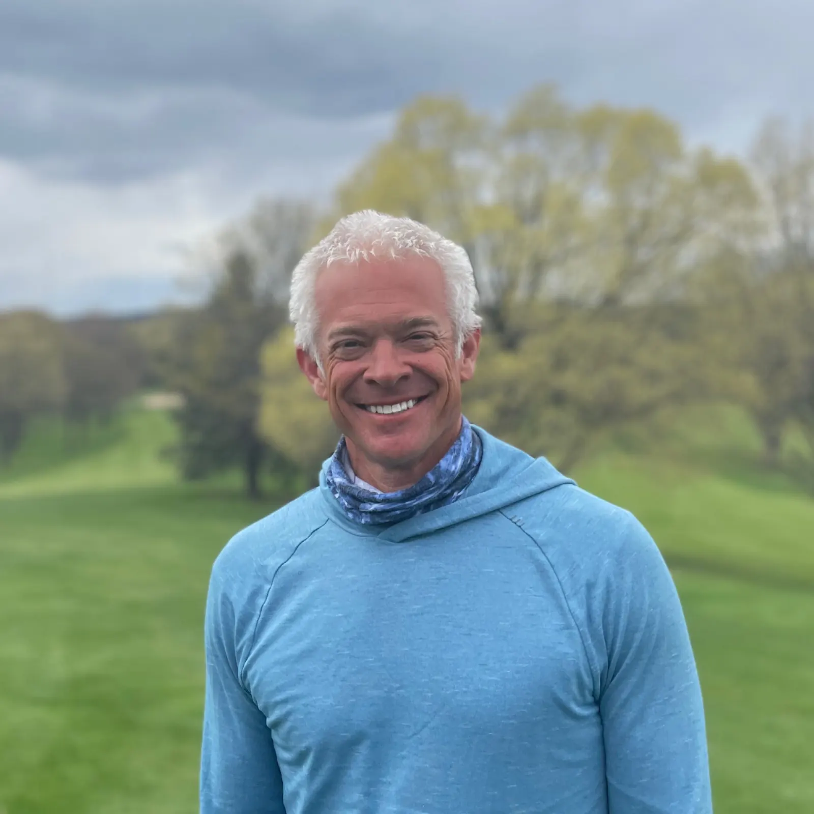 Derek Schnarr smiling outdoors in a blue hooded top with a golf fairway blurred behind him