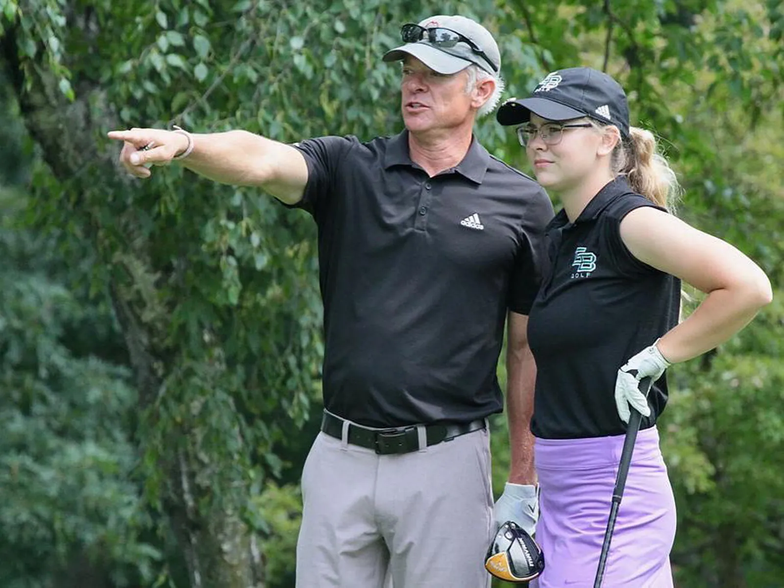 Derek Schnarr points down the hole beside a junior golfer holding a driver, with trees behind them