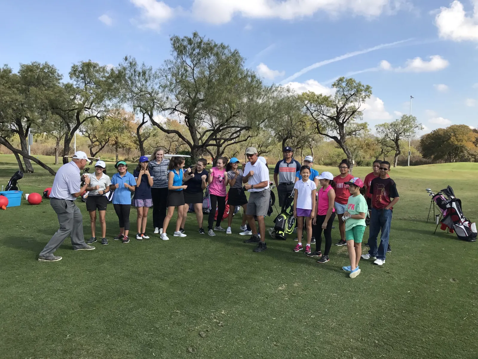 Group of young golfers and two adults standing on a grass fairway lined with trees, junior golf program