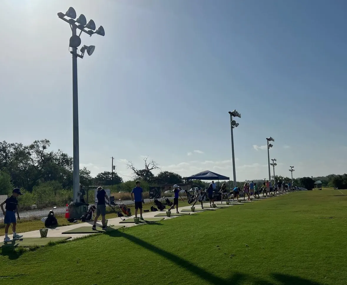 Row of young golfers swinging along the hitting line under floodlight poles, David Ogrin Golf Academy driving range
